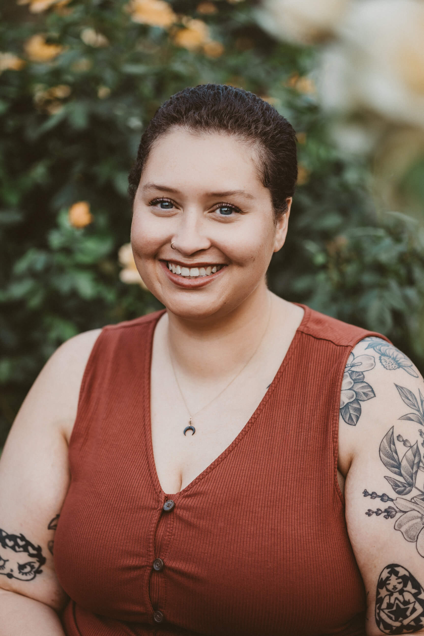 A headshot photo of Sophia Starks-Bailey, the 11th House Doula. She is looking at the camera, smiling. She is wearing a burnt orange dress and sitting in front of a Rose Garden. 