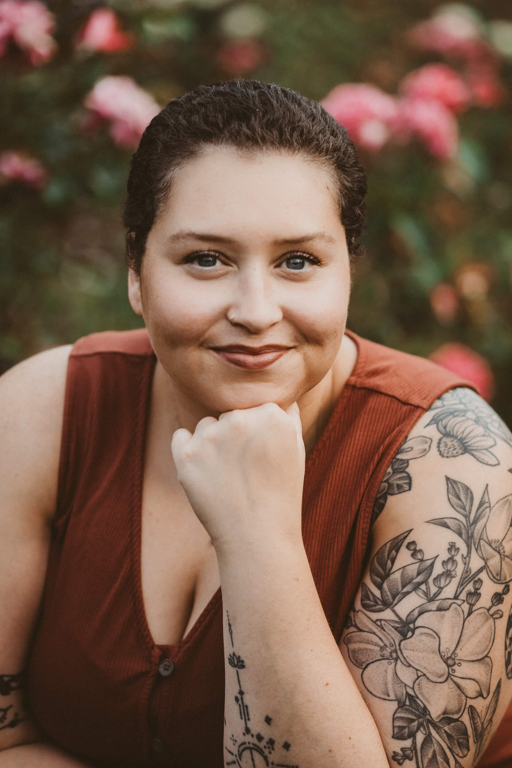 A photo of Sophia Starks-Bailey, the owner of 11th House Doula Services. They are smiling, looking at the camera with their chin resting on their hand. They are wearing an orange top and photographed in a Portland Rose Garden. 